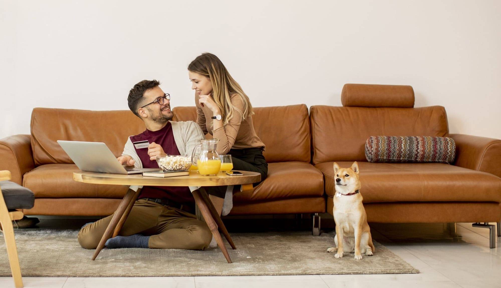 casal feliz na sala de estar, com seu cachorro. Um notebook sobre a mesa, pote com pipocas, suco de laranja.